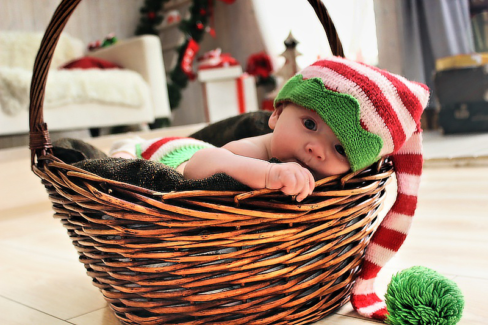 Baba fotó retusálás 04 - Pár napos baba egy fonott kosárból integet kifelé a fején egy piros-fehér-zöld bojtos végű sapkában.