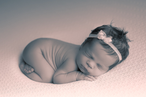 Baba fotó retusálás 07 - Alvó újszülött baba süpped bele az ágy szivacsába egy fehér csipkés hajpánttal a fején.