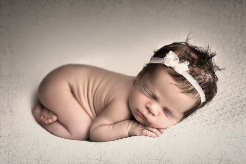 Baba fotó retusálás 08 - Alvó újszülött baba süpped bele az ágy szivacsába egy fehér csipkés hajpánttal a fején.
