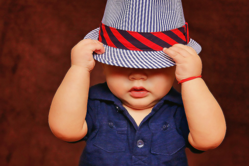 Baba fotó retusálás 16 - Fiú baba kék ingben, fehér-kék csíkos sapkában, és a sapkán egy ferde-csíkozású, piros-fekete színes sapkában. A kalapot a kezében tartja és az arcát félig eltakarja.