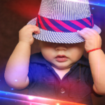 Baba fotó retusálás 17 - Fiú baba kék ingben, fehér-kék csíkos sapkában, és a sapkán egy ferde-csíkozású, piros-fekete színes sapkában. A kalapot a kezében tartja és az arcát félig eltakarja.