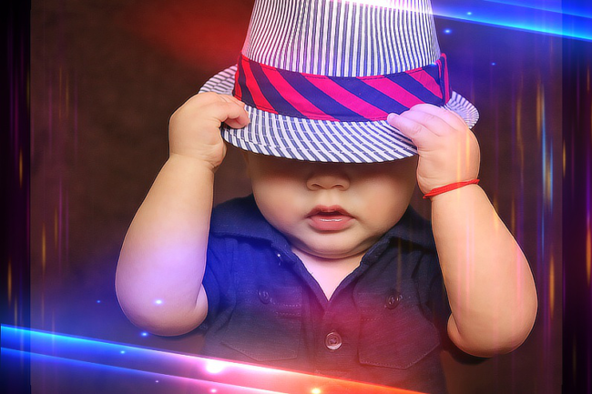 Baba fotó retusálás 17 - Fiú baba kék ingben, fehér-kék csíkos sapkában, és a sapkán egy ferde-csíkozású, piros-fekete színes sapkában. A kalapot a kezében tartja és az arcát félig eltakarja.
