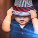 Baba fotó retusálás 18 - Fiú baba kék ingben, fehér-kék csíkos sapkában, és a sapkán egy ferde-csíkozású, piros-fekete színes sapkában. A kalapot a kezében tartja és az arcát félig eltakarja.